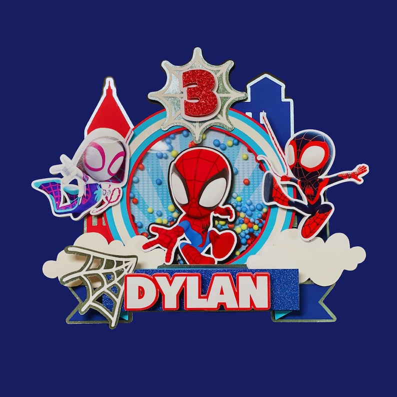 May include: A colorful cake topper featuring Spider-Man characters. The design includes the number 3, the name "Dylan", and various superhero elements. The background is a deep blue.