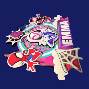 May include: A colorful cake topper featuring Spider-Man and other superhero-themed elements. The topper includes a pink and white circular design with a clear center filled with confetti, and the name "EMMA" in purple glitter.