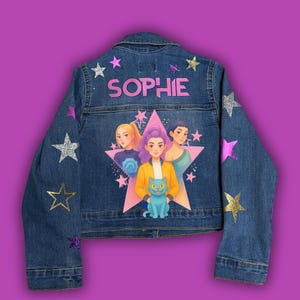 Puede incluir: Una chaqueta de mezclilla azul con el nombre SOPHIE en letras rosas en la parte superior. La espalda muestra un gráfico de estrella rosa grande con tres personajes ilustrados y un gato azul. La chaqueta está decorada con varias estrellas brillantes en rosa, plata y oro.