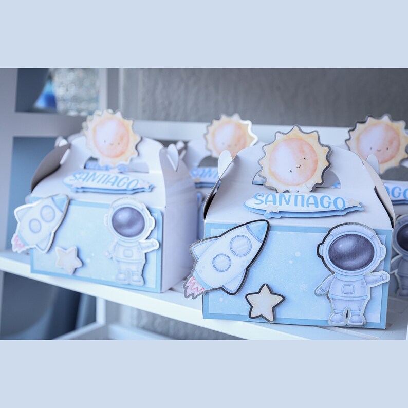 May include: Light blue favor boxes decorated with space-themed cutouts, including rockets, astronauts, and stars. Each box has a handle and a sun-shaped topper. The name "SANTIAGO" is printed on each box.
