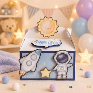 May include: A white gable box with a blue and white space theme. The box features a smiling sun, a rocket, an astronaut, and a star. The name "Zayne Apollo" is written on the box. The box is decorated with blue and purple balloons.