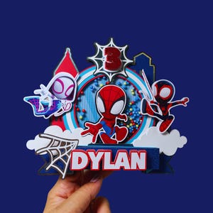 May include: A colorful cake topper featuring Spider-Man characters. The design includes Spider-Man, Spider-Gwen, and Miles Morales, with a number 3 and the name "Dylan". The background is a blue and white circular design with a blue base.