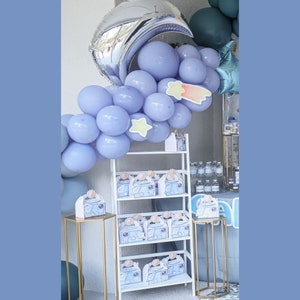 May include: A party decoration setup featuring a celestial theme. A silver crescent moon balloon and light blue balloons form an arch. A white shelf displays gift boxes with a space-themed design. Gold stands hold additional gift boxes.