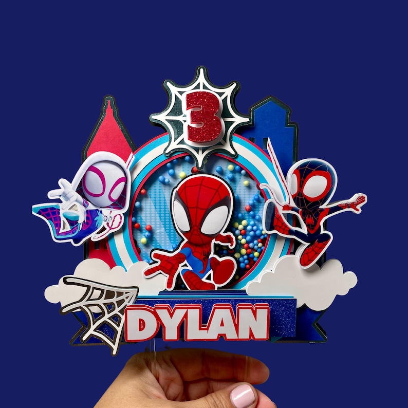 May include: A colorful cake topper featuring Spider-Man characters. The design includes Spider-Man, Spider-Gwen, and Miles Morales, with a glittery number 3 and the name "Dylan". The background is a vibrant blue.
