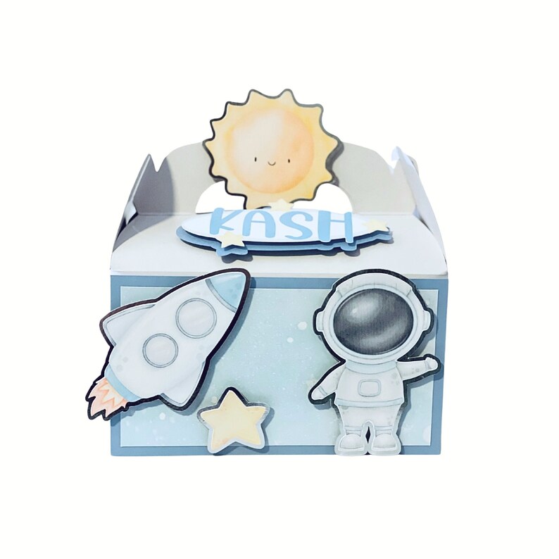 May include: A light blue and white gift box with a space theme. The box features a rocket, astronaut, star, and a smiling sun. The name "KASH" is written in blue on a blue banner. The box is perfect for a space-themed party.