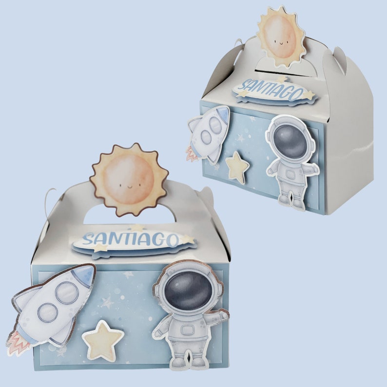 May include: Two white favor boxes with a space theme. Each box features a blue background with a rocket, astronaut, and star stickers. The boxes have a sun-shaped handle and the name "SANTIAGO" on the front.