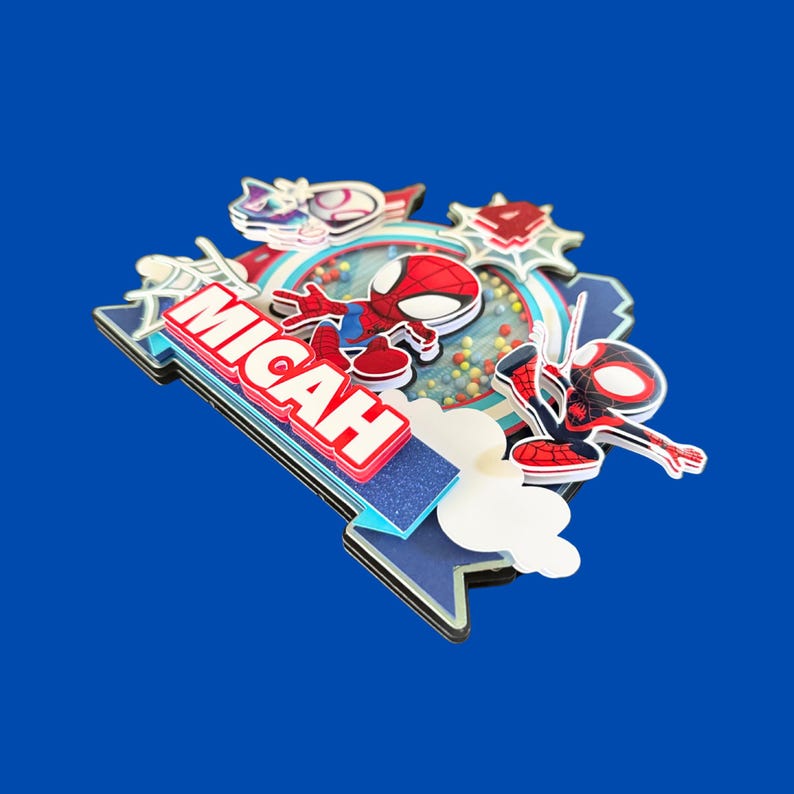 May include: A multi-layered cake topper featuring Spider-Man characters. The topper includes the name "MICAH" in red and white, along with various decorative elements and a blue background.