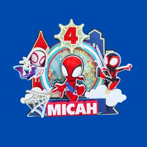 May include: A colorful cake topper featuring Spider-Man and other characters. The number "4" is displayed, along with the name "MICAH". The design includes a cityscape and a web, all set against a blue background.