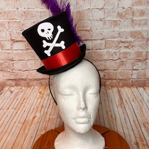 May include: A black top hat with a red band and a white skull and crossbones design. The hat is adorned with a purple feather and is attached to a black headband. The hat is displayed on a white mannequin head.