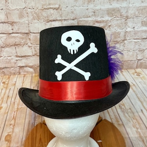 May include: Black top hat with a red band and a white skull and crossbones design. A purple feather is attached to the side of the hat.
