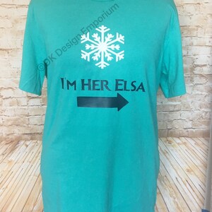 May include: Teal blue t-shirt with a white snowflake and black text that reads "I'm Her Elsa" with a black arrow pointing to the right.