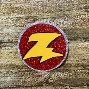 May include: A red glitter circle with a silver border and a yellow lightning bolt shaped "Z" in the center.