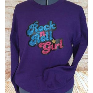 May include: Purple sweatshirt with the text "Rock n Roll Girl" in blue and pink glitter lettering.
