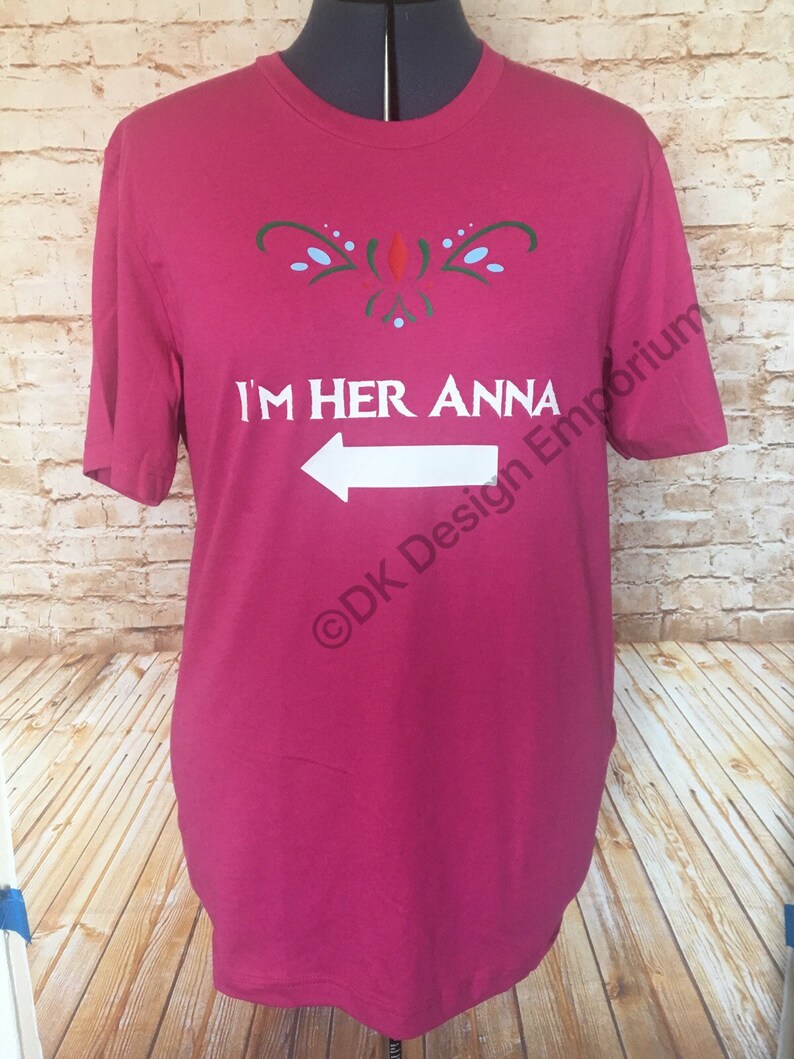 May include: A pink t-shirt with a white arrow pointing to the left and the text "I'm Her Anna" in white. The shirt has a green and blue design above the text. The text "&copy;DK Design Emporium" is in the bottom right corner.