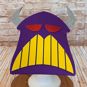 May include: Purple monster head costume with yellow teeth and red eyes. The costume is made of felt and has a white base.