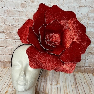 May include: A large, red glitter flower headpiece. The flower is attached to a black headband and is displayed on a white mannequin head. The flower has multiple layers of petals and a detailed center.