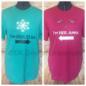 May include: Two t-shirts, one turquoise and one pink, with a snowflake and a stylized floral design. The turquoise shirt says "I'm her Elsa" with a black arrow pointing right. The pink shirt says "I'm her Anna" with a white arrow pointing left.