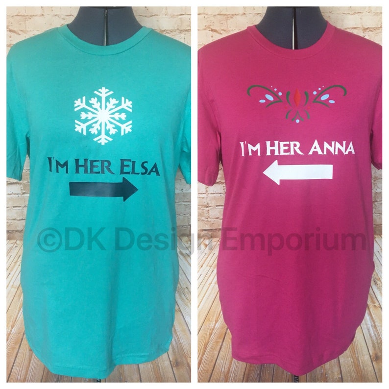 May include: Two t-shirts, one turquoise and one pink, with a snowflake and a stylized floral design. The turquoise shirt says "I'm her Elsa" with a black arrow pointing right. The pink shirt says "I'm her Anna" with a white arrow pointing left.