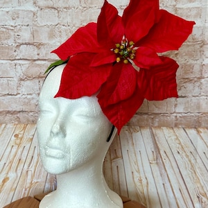 May include: A festive red poinsettia flower headpiece. The large, vibrant red petals are accented with small yellow and green details at the center. The flower is attached to a black headband, perfect for holiday celebrations.