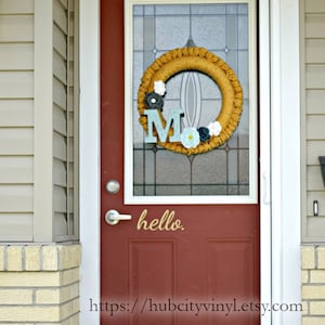 May include: A red front door with a glass panel and a crocheted wreath featuring the letter "M" and flowers. The door has a gold vinyl decal that says "hello" and a website address at the bottom.
