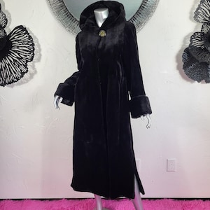 May include: A long, black faux fur coat with a hood and cuff details. The coat features a gold-colored button closure and a side slit. The coat is displayed on a mannequin.