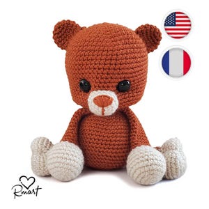 May include: A crocheted brown teddy bear with white paws sitting on a white background. The bear has black eyes and a white nose.