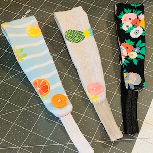 May include: Three fabric headbands with button closures. One headband is light blue with a citrus fruit pattern, another is gray with a pineapple design, and the last is black with a floral pattern.