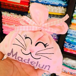 May include: A pink plush bunny-shaped bag with a pink and white gingham ribbon. The bag has an embroidered smiling face with whiskers and the name "Madelyn" on the front.