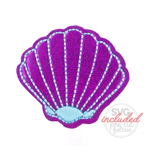 Può includere: Una conchiglia di glitter viola con un interno blu chiaro. La conchiglia è bordata con filo bianco.
