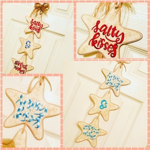 Puede incluir: Un conjunto de tres adornos de estrella de mar beige con las palabras "Salty Kisses" y "Starfish Wishes" bordadas en hilo azul y rojo. Los adornos están ensartados con un cordel y están listos para colgar.