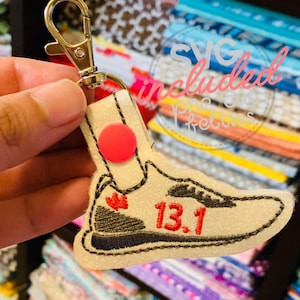 May include: A white and black running shoe keychain with red glitter and the number "13.1" embroidered on it. The keychain has a silver metal key ring.