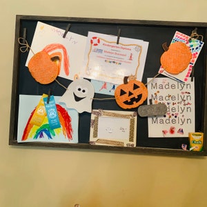 Può includere: Una lavagna nera con una varietà di decorazioni, tra cui un disegno di arcobaleno, un fantasma, una zucca, un diploma e un nome scritto con lettere colorate. La lavagna include anche una piccola scatola di gesso.