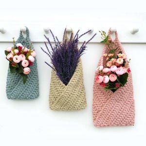 May include: Three crocheted hanging planters with different colored yarn. The planters are filled with artificial flowers. The planters are hanging from a white wall.