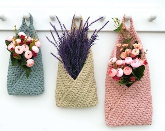 May include: Three crocheted hanging planters with different colored yarn. The planters are filled with artificial flowers. The planters are hanging from a white wall.