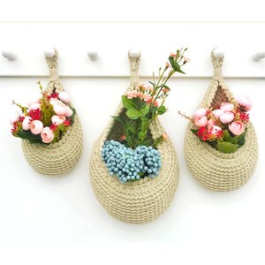 May include: Three hanging macrame plant hangers with a teardrop shape. The hangers are made of natural beige rope and are filled with artificial flowers. The flowers are pink, white, and blue.