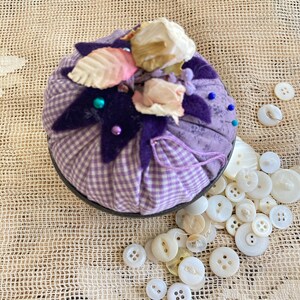 May include: A round, purple and white gingham pin cushion with dried flowers and decorative beads. A collection of white buttons of various sizes are scattered around the pin cushion on a textured, beige surface.