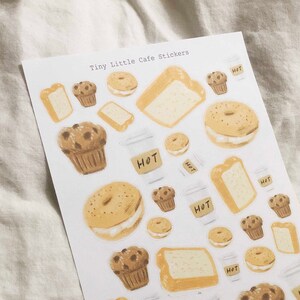 May include: A sheet of stickers featuring illustrations of various bakery items, including muffins, bagels, bread, and coffee cups. The stickers are white with a light brown background and have the text "Tiny Little Cafe Stickers" and "Pearlteapaperie" printed on them.