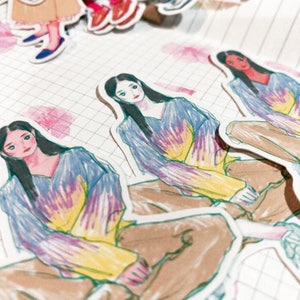 May include: A collection of stickers featuring illustrations of a person in a tie-dye top and brown pants. The stickers are on a grid-lined paper with pink floral accents. The person has long black hair.