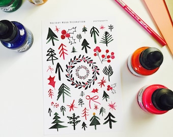 May include: A white paper with watercolor illustrations of Christmas trees, holly, berries, and wreaths. The words "Holiday Mood Decoration" are printed at the top. Ink bottles and a red pencil are arranged around the paper.