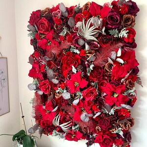 May include: A rectangular wall hanging densely packed with red roses, poinsettias, and other flowers. Accents include silver-gray leaves and red feathers. The arrangement is a vibrant display of floral artistry.