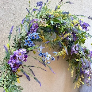 May include: A colourful floral archway arrangement with purple, blue, and yellow flowers and green leaves. The flowers include lavender and orchids. Ideal for weddings or home decoration. The arrangement is in an arch shape.