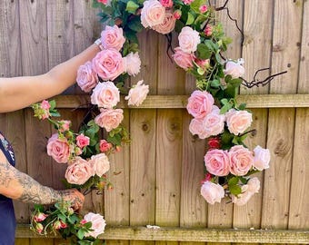 Puede incluir: Una guirnalda floral rosa y verde con rosas y vegetación, colgada de una valla de madera.