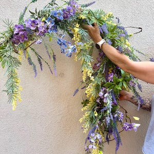 May include: A floral arrangement featuring a variety of artificial flowers in shades of purple, blue, and yellow. The arrangement includes lavender, orchids, and other greenery, creating a natural and vibrant look. The flowers are arranged in a cascading style.