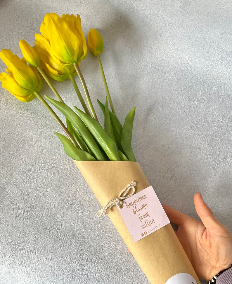 May include: A bouquet of yellow tulips wrapped in brown paper with a tag that reads "happiness blooms from within".