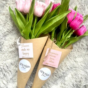 Op de afbeelding: Twee boeketten tulpen verpakt in bruin papier. E&eacute;n boeket heeft lichtroze tulpen, de andere levendige fuchsia tulpen. Elk boeket heeft een label met de tekst "Flowers make me happy" en "happiness blooms from within", samen met het "SDesign Floral" logo.
