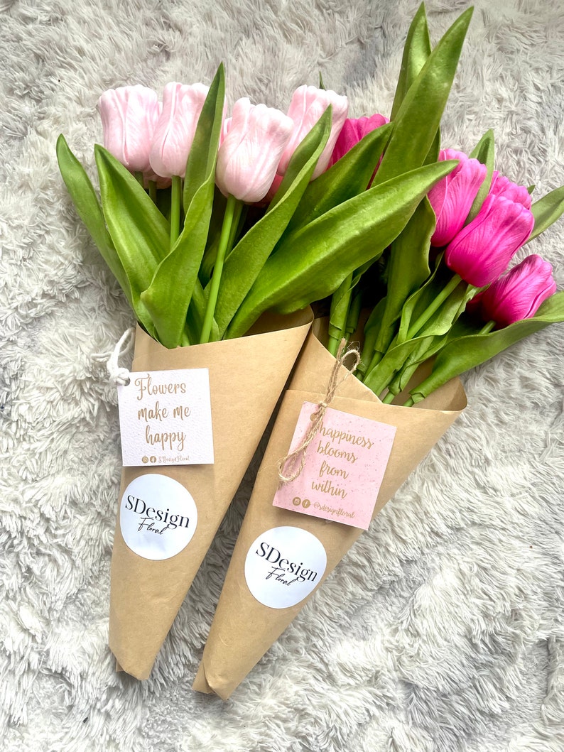Op de afbeelding: Twee boeketten tulpen verpakt in bruin papier. E&eacute;n boeket heeft lichtroze tulpen, de andere levendige fuchsia tulpen. Elk boeket heeft een label met de tekst "Flowers make me happy" en "happiness blooms from within", samen met het "SDesign Floral" logo.