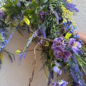 May include: A vibrant floral arrangement featuring purple orchids, lavender, and yellow mimosa. Green leaves and brown branches add texture. The arrangement is a mix of artificial and natural elements, creating a colorful and lively display.