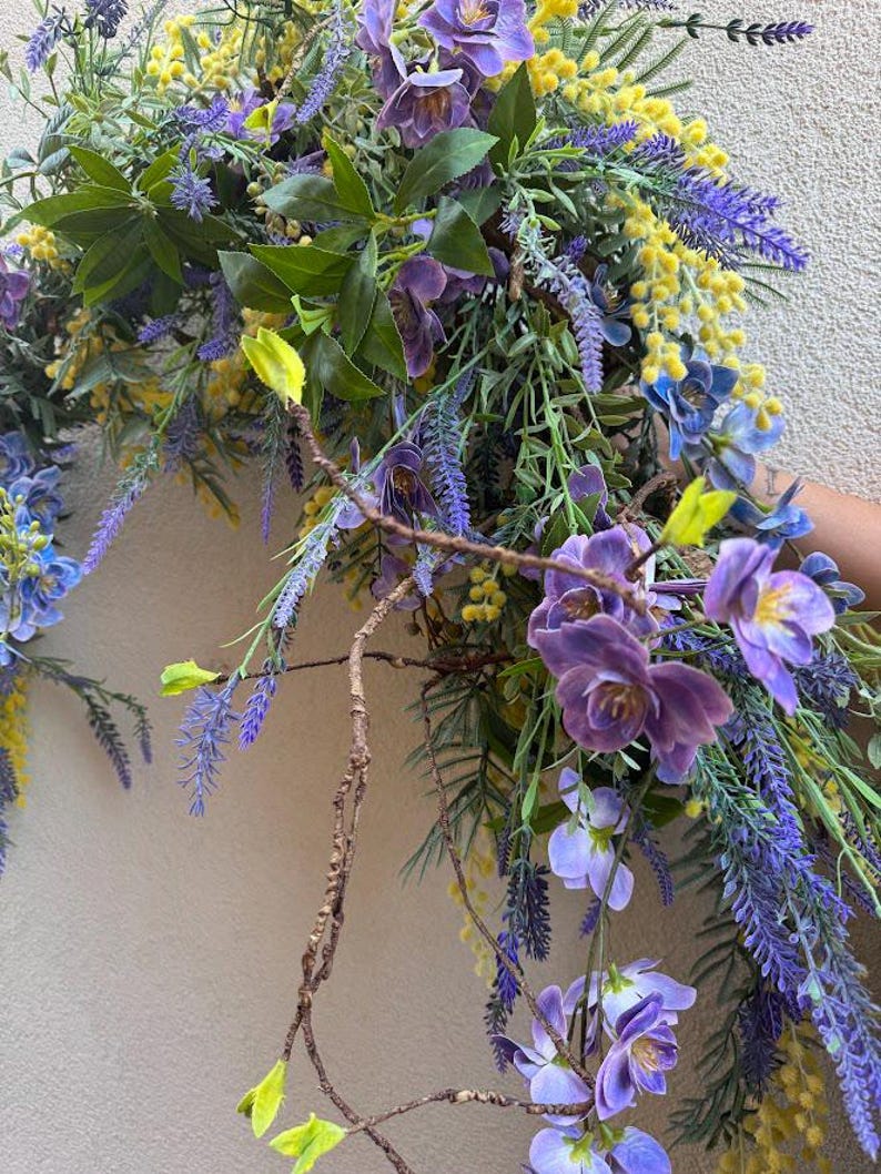 May include: A vibrant floral arrangement featuring purple orchids, lavender, and yellow mimosa. Green leaves and brown branches add texture. The arrangement is a mix of artificial and natural elements, creating a colorful and lively display.