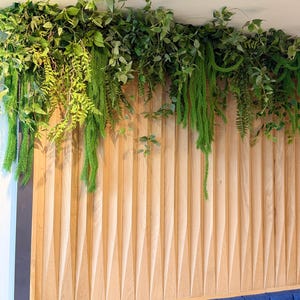 May include: A decorative wall featuring a wooden panel with a geometric design, adorned with lush green foliage. Below, a blue upholstered bench provides a contrasting color. A gold-colored spotlight is mounted on the ceiling.