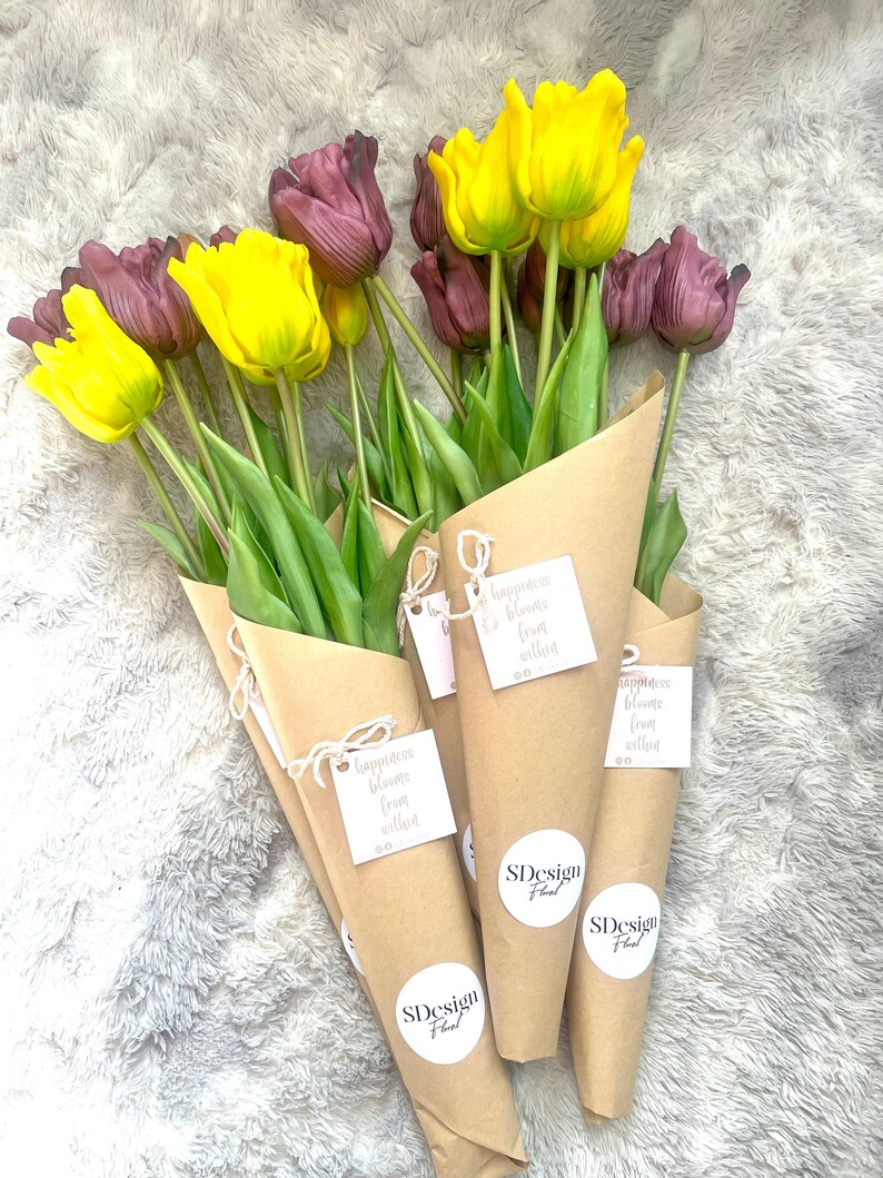 May include: A bouquet of yellow and purple tulips wrapped in brown paper with a tag that reads "Happiness blooms from within". The bouquet is tied with twine and has a round sticker that says "SDesign Floral".